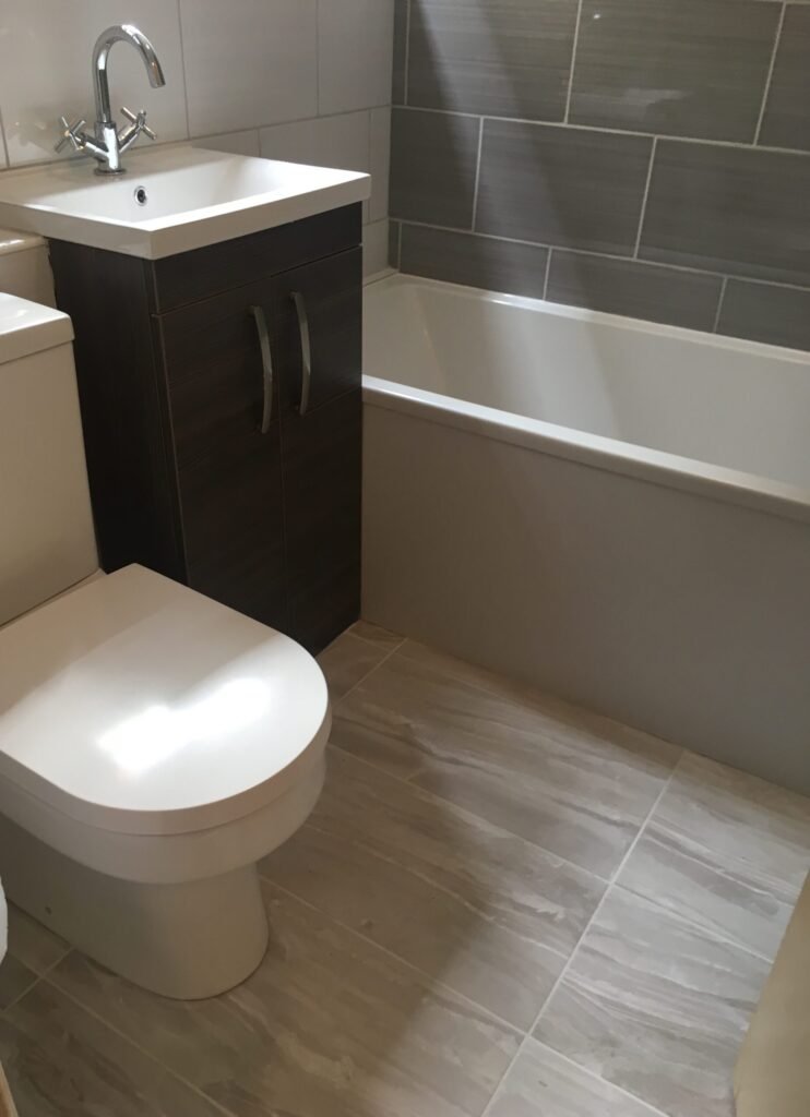 "Modern bathroom installation with grey tiles, white bathtub, dark wood vanity unit, and white toilet"